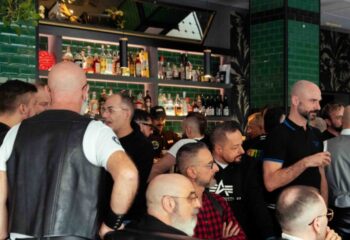 Leather Club Roma Event: Gay men gather at a bar. Some wear leather vests. Bottles line the shelves behind the counter. Nightlife scene.