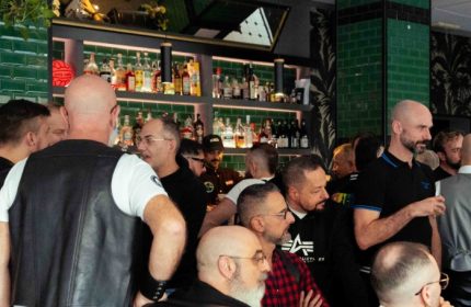 Leather Club Roma Event: Gay men gather at a bar. Some wear leather vests. Bottles line the shelves behind the counter. Nightlife scene.