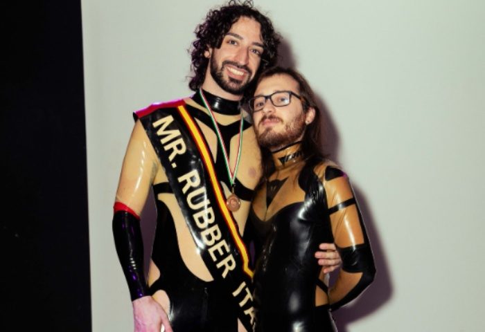 Leather Club Roma Event: Two gay men in latex outfits. One wears a sash reading Mr. Rubber Italia at a fetish event.