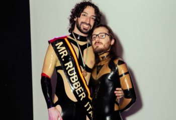 Leather Club Roma Event: Two gay men in latex outfits. One wears a sash reading Mr. Rubber Italia at a fetish event.