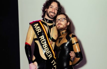 Leather Club Roma Event: Two gay men in latex outfits. One wears a sash reading Mr. Rubber Italia at a fetish event.