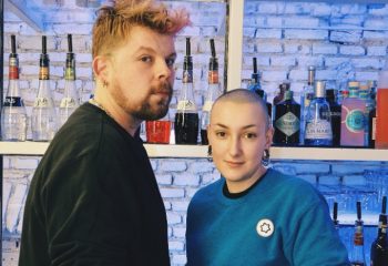 Lecco, Milan: Two stylish people at a bar. Bottles of liquor line the shelves behind them. Gay travel advice for Italy.