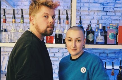 Lecco, Milan: Two stylish people at a bar. Bottles of liquor line the shelves behind them. Gay travel advice for Italy.