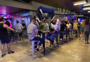 Lights Bar, a gay bar in Torremolinos, Spain. People socialize at tables, some wearing masks. Balloons decorate the entrance. Nightlife scene.