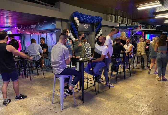 Lights Bar, a gay bar in Torremolinos, Spain. People socialize at tables, some wearing masks. Balloons decorate the entrance. Nightlife scene.