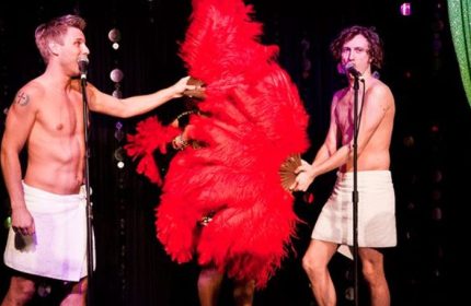 Lips Drag Queen Show Palace in New York: Two shirtless men in towels flank a drag queen with a large red feather fan, performing on stage.