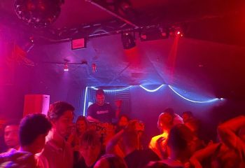Lola's Club Ibiza gay club: DJ playing music to a crowd of people under red and blue lights. Nightlife scene in Ibiza for gay men.