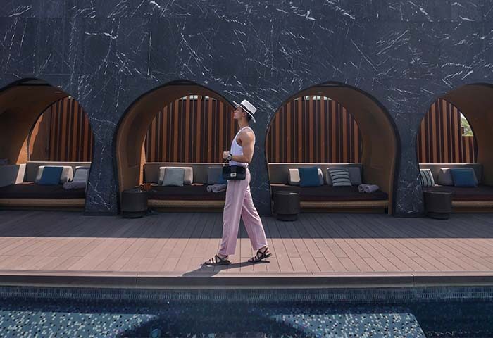 Gay traveler at a luxe, gay-friendly hotel in Chiang Mai. Man in hat walks by the pool, with cabanas in the background. Thailand travel.