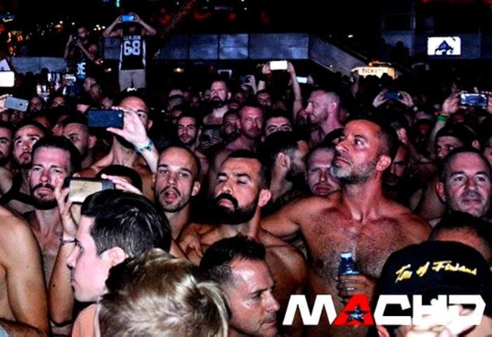 Crowd at MACHO gay party in Madrid. Many shirtless men, some taking photos. Nightlife scene, popular gay club.
