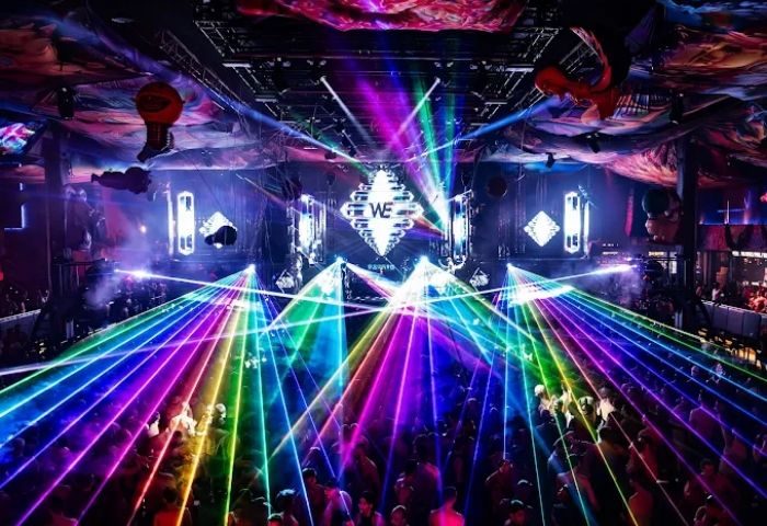 Madrid's MACHO Party at a gay club. Crowd with rainbow lasers, stage lights, and WE logo. Vibrant nightlife for gay men travel.