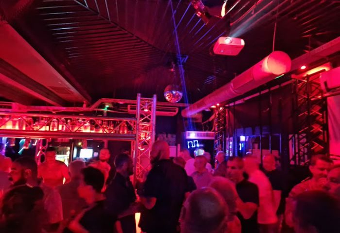 Gay cruise club in Torremolinos. Men gather under red lights, a disco ball, and a projector. Nightlife scene for gay men travelers.