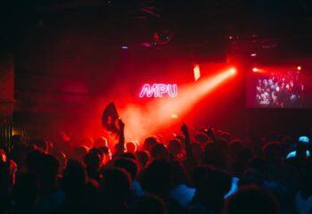 Montreal gay nightlife: Le Belmont's MPU dance party. Crowd with raised hands, red lighting, and a neon MPU sign. Nightclub scene.