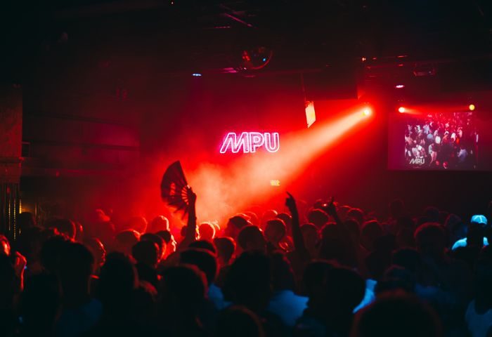 Montreal gay nightlife: Le Belmont's MPU dance party. Crowd with raised hands, red lighting, and a neon MPU sign. Nightclub scene.