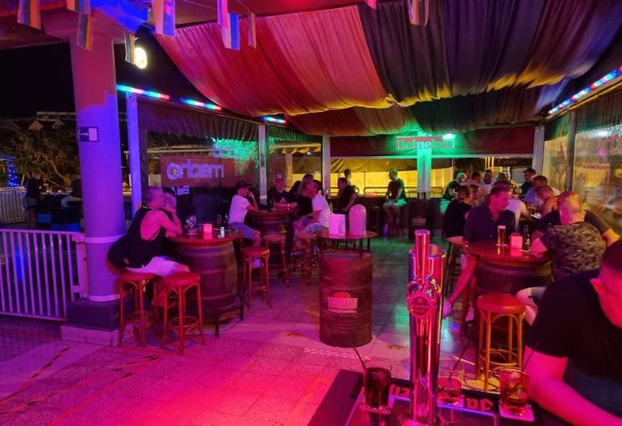 Macho Macho Gay Bar in Gran Canaria: Interior view of the bar, patrons sit at tables made from barrels, enjoying drinks. Colorful lights.
