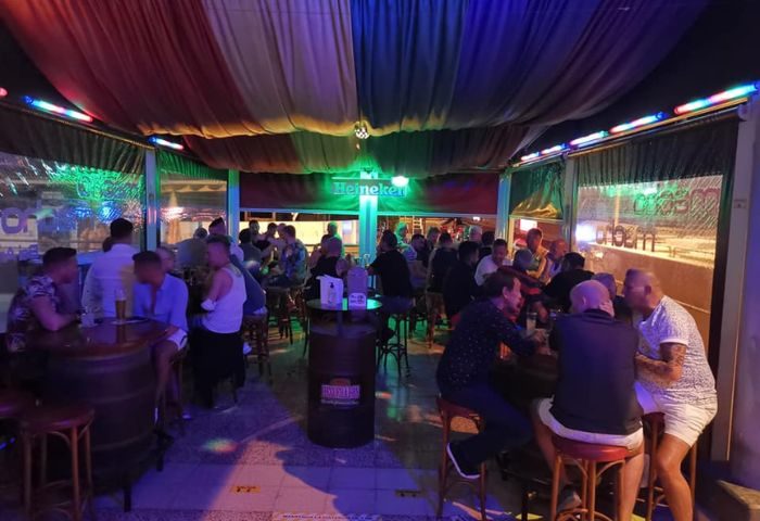 Interior of a gay bar in Gran Canaria. Men are at tables and the bar. The ceiling is draped with rainbow fabric. Macho Macho Bar.