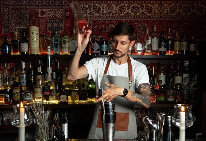 Bartender making a cocktail at Madame George Lounge Bar, a gay bar in Barcelona. Bottles line the shelves in the background. Gay travel.