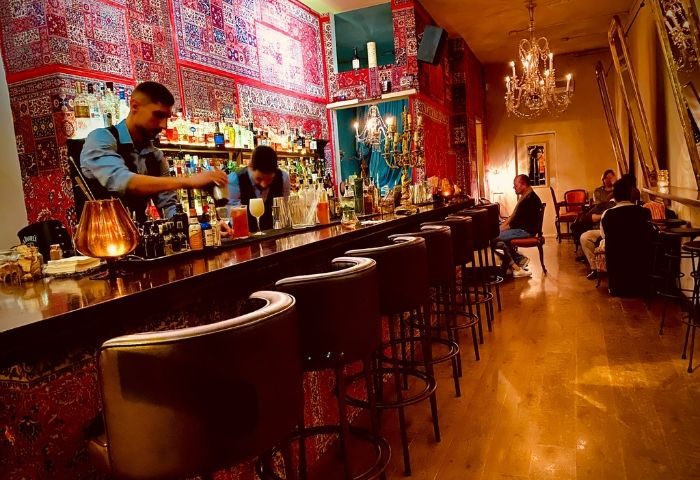 Madame George Lounge Bar, a gay bar in Barcelona. Bartenders mix drinks at a long bar with red patterned walls and chandelier lighting.