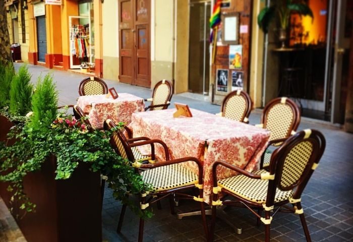 Outdoor seating at Madame George Lounge Bar, a gay bar in Barcelona. Tables with patterned cloths and wicker chairs line the street.