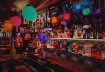 Madame Jasmine Gay Bar in Barcelona, Spain. Shelves of liquor bottles behind the bar with colorful lighting. Gay travel advice.