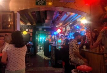 Interior view of Madame Jasmine Gay Bar in Barcelona. People at bar, colorful lights, and exit sign. Gay travel advice.