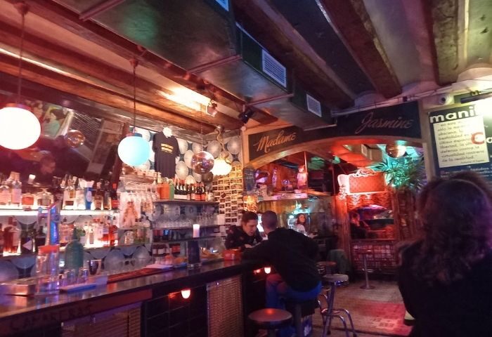 Madame Jasmine Gay Bar interior, Barcelona. A cozy, dimly lit space with a well-stocked bar and patrons enjoying drinks. Gay travel advice.