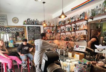 Magnebevo bar at Pigneto, Rome. Gay travel: interior view with shelves of liquor, barman, and patrons. Rainbow flag visible.