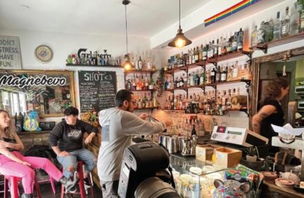 Magnebevo bar at Pigneto, Rome. Gay travel: interior view with shelves of liquor, barman, and patrons. Rainbow flag visible.