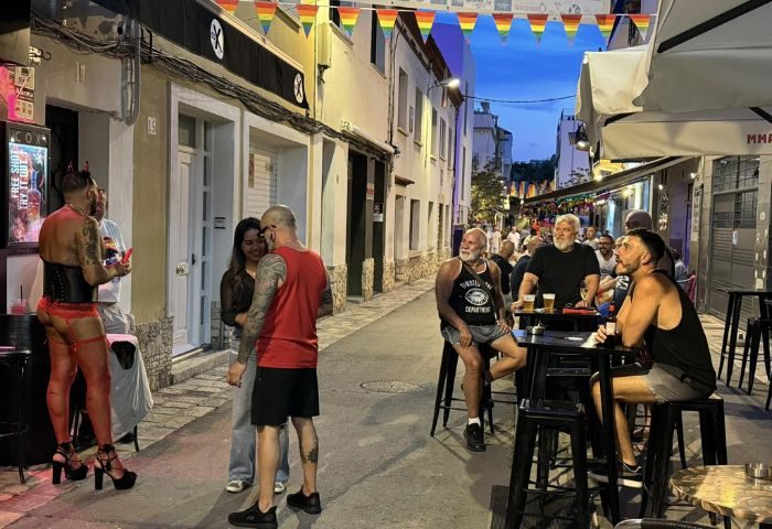 Sitges gay bar scene at night. Men gather, some at tables, others near a bar with a performer in red lingerie and high heels. Rainbow flags above.