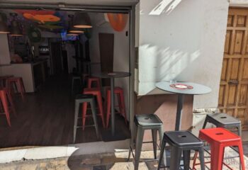 Sitges gay bar entrance. Red and gray stools line the sidewalk and interior. MAN sign visible inside. Gay travel advice.