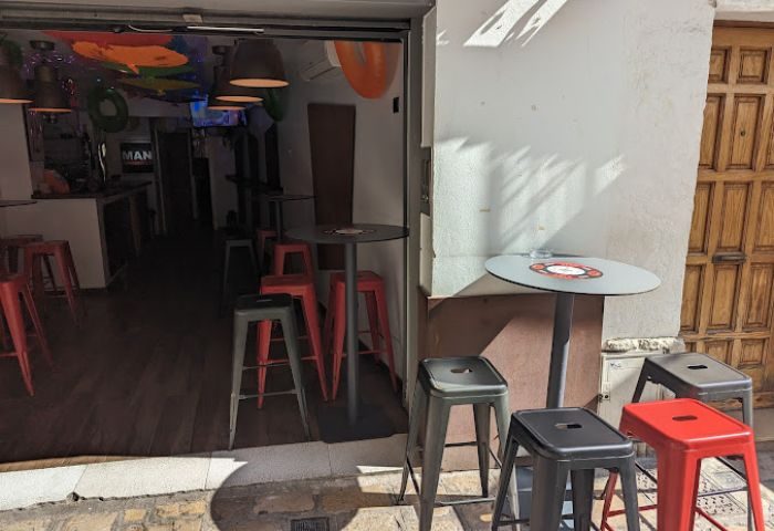 Sitges gay bar entrance. Red and gray stools line the sidewalk and interior. MAN sign visible inside. Gay travel advice.