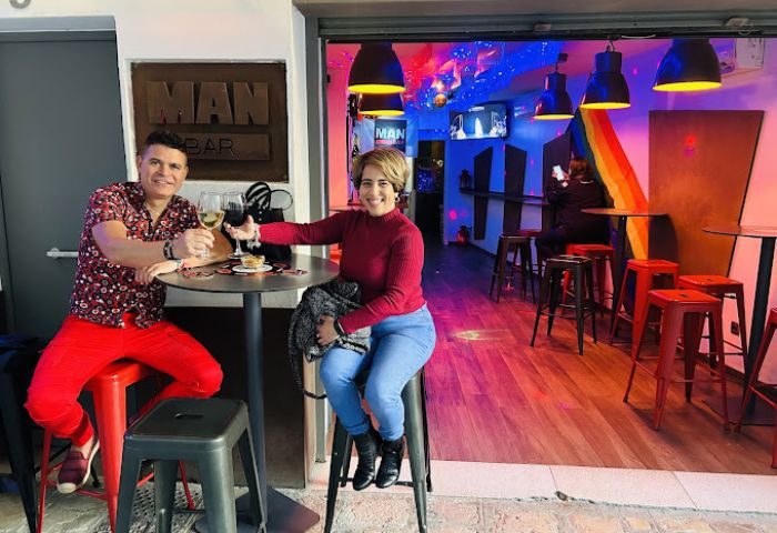 Couple at Man Bar, a gay bar in Sitges, Spain. They're drinking wine. The bar has rainbow decor and a lively atmosphere. Gay travel destination.