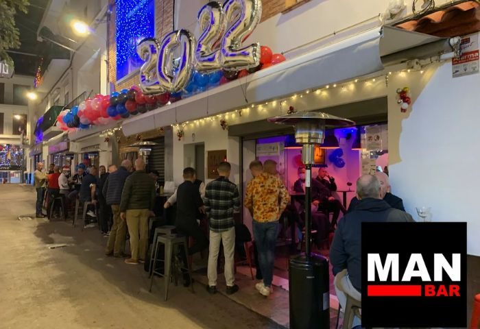 Man Bar in Sitges, a popular gay bar, is crowded with men outside. Balloons and lights decorate the entrance. A vibrant nightlife scene.