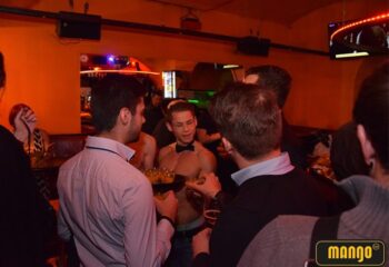 Inside Mango Bar Vienna: A shirtless waiter with a bow tie serves drinks to gay men. Nightlife scene at a gay bar.