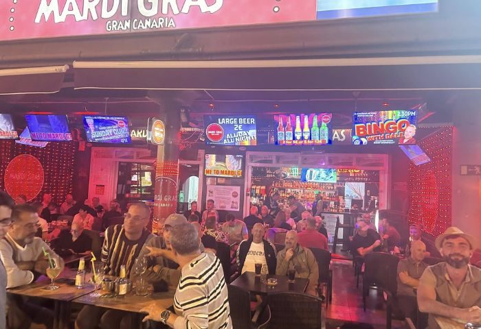 Mardi Gras Gay Bar in Gran Canaria. Interior view of a busy gay bar with men at tables, TVs showing ads, and a well-stocked bar in the back.