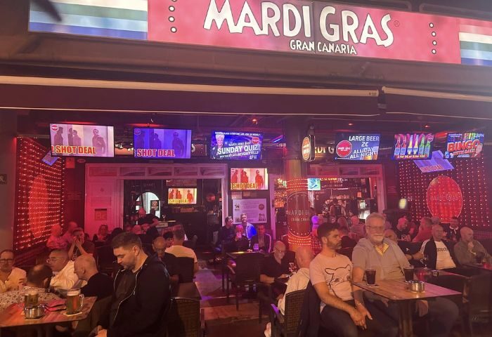 Mardi Gras gay bar in Gran Canaria, a popular gay travel destination. Men are seated at tables, enjoying drinks under TV screens. Nightlife scene.