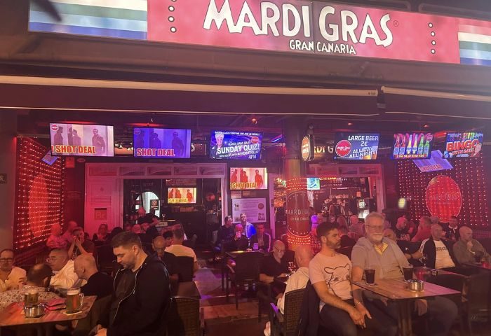 Mardi Gras gay bar in Gran Canaria, a popular gay travel destination. Men are seated at tables, enjoying drinks under TV screens. Nightlife scene.