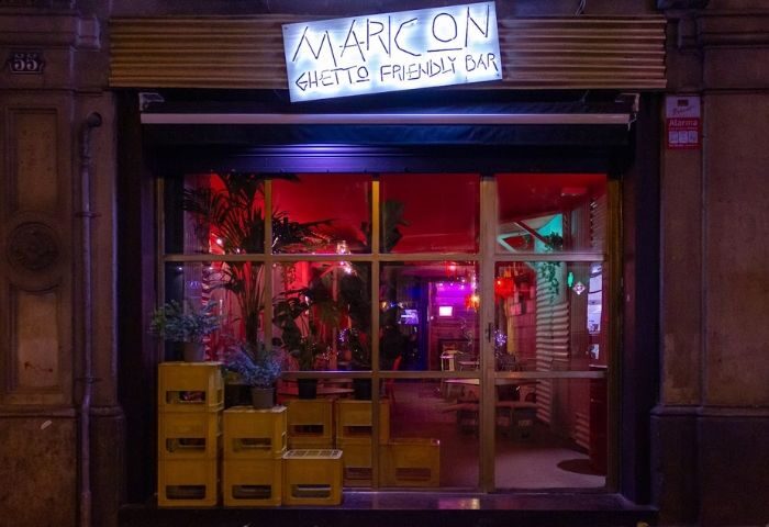 Barcelona's Maricon gay bar facade. Ghetto-friendly vibe with plants, yellow crates, and neon sign. A unique spot in Barcelona's gay scene.