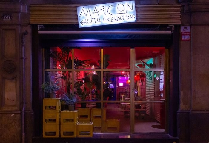 Barcelona's Maricon gay bar facade. Ghetto-friendly vibe with plants, yellow crates, and neon sign. A unique spot in Barcelona's gay scene.