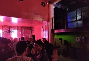 Interior of Marrakech Salon Gay Bar in Mexico. People socializing, bar with bottles, red lighting, and a mezzanine level. Gay travel advice.