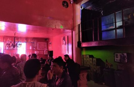 Interior of Marrakech Salon Gay Bar in Mexico. People socializing, bar with bottles, red lighting, and a mezzanine level. Gay travel advice.