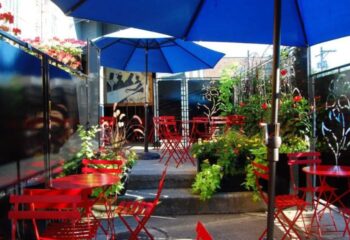 Outdoor patio at Marty's Martini Bar, a gay bar in Chicago. Red chairs and tables under blue umbrellas, plants, and decorative screens.