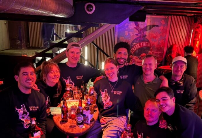 Group at Masken Bar, Copenhagen. Gay travel advice: People smiling, wearing Masken Bar shirts, drinks on table. Nightlife scene.