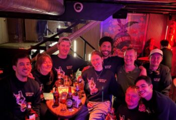 Group at Masken Bar, Copenhagen. Gay travel advice: People smiling, wearing Masken Bar shirts, drinks on table. Nightlife scene.