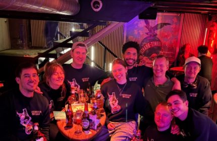 Group at Masken Bar, Copenhagen. Gay travel advice: People smiling, wearing Masken Bar shirts, drinks on table. Nightlife scene.