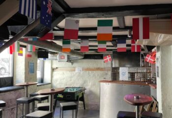 Masken Bar Copenhagen interior. Flags hang from the ceiling. Foosball table, bar stools, and tables. Gay travel advice.