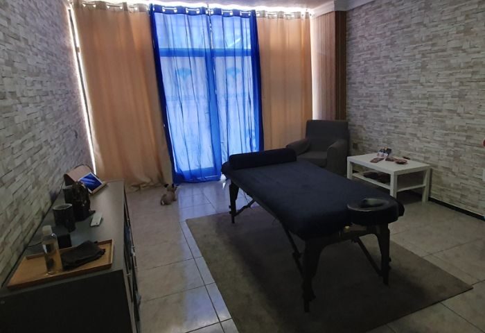 Gay Maspalomas: Massage room in Gran Canaria, featuring a massage table, chair, and a small dog near the window. Cozy spa setting.