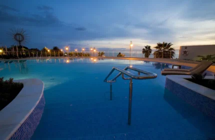 Meliá Madeira Mare Hotel pool at dusk. Gay travel in Funchal, Portugal. Relaxing vacation spot with ocean views. Palm trees and lounge chairs.
