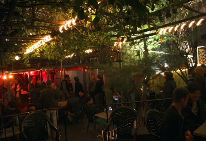 Outdoor area of Metropolitan Bar for queers in New York. People at tables and standing, with lights and greenery overhead. Gay bar scene.