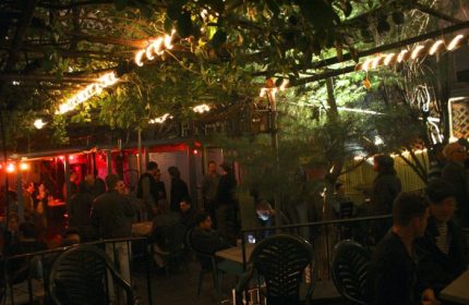 Outdoor area of Metropolitan Bar for queers in New York. People at tables and standing, with lights and greenery overhead. Gay bar scene.