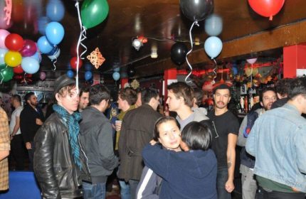 Inside Metropolitan Bar for Queers in New York, a lively crowd of gay men mingle beneath colorful balloons, enjoying drinks & camaraderie.